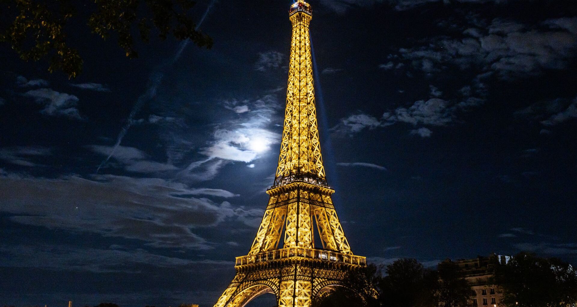 A view looking up at the Eiffel Tower lit in gold at night shining above the Paris skyline as the moon illuminates a bank of dappled clouds to the left of the steel structure.