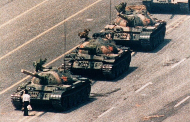A lone person stands in front of a line of three military tanks on a wide, empty street, blocking their path.
