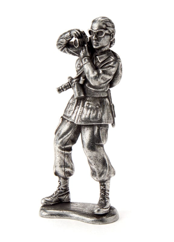 A silver metal figurine of a person in military attire stands upright, holding binoculars to their eyes. They wear boots, a backpack, and have a sword at their side. The figure is detailed and photographed on a white background.