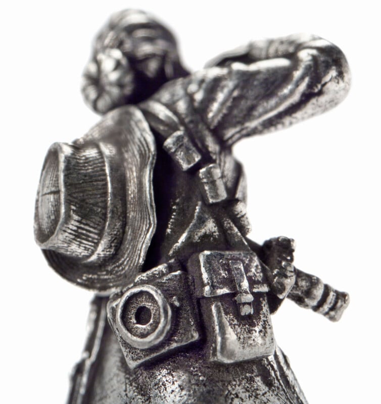 A close-up of a silver figurine of an explorer wearing a hat and backpack, with binoculars and a canteen strapped to their belt, looking off into the distance. The background is white.