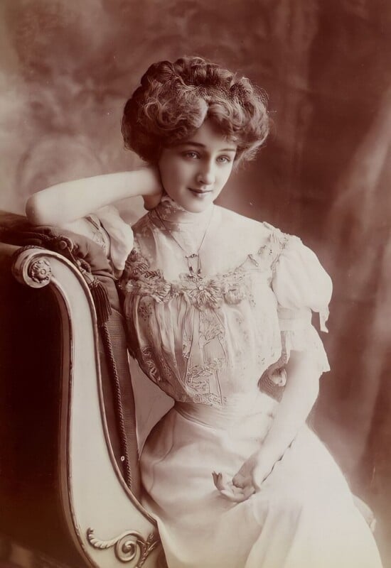 A young woman in an elegant, high-necked Edwardian dress sits on an ornate chair, resting her head on her hand and smiling softly. The sepia-toned photo gives a vintage, early 20th-century feel.