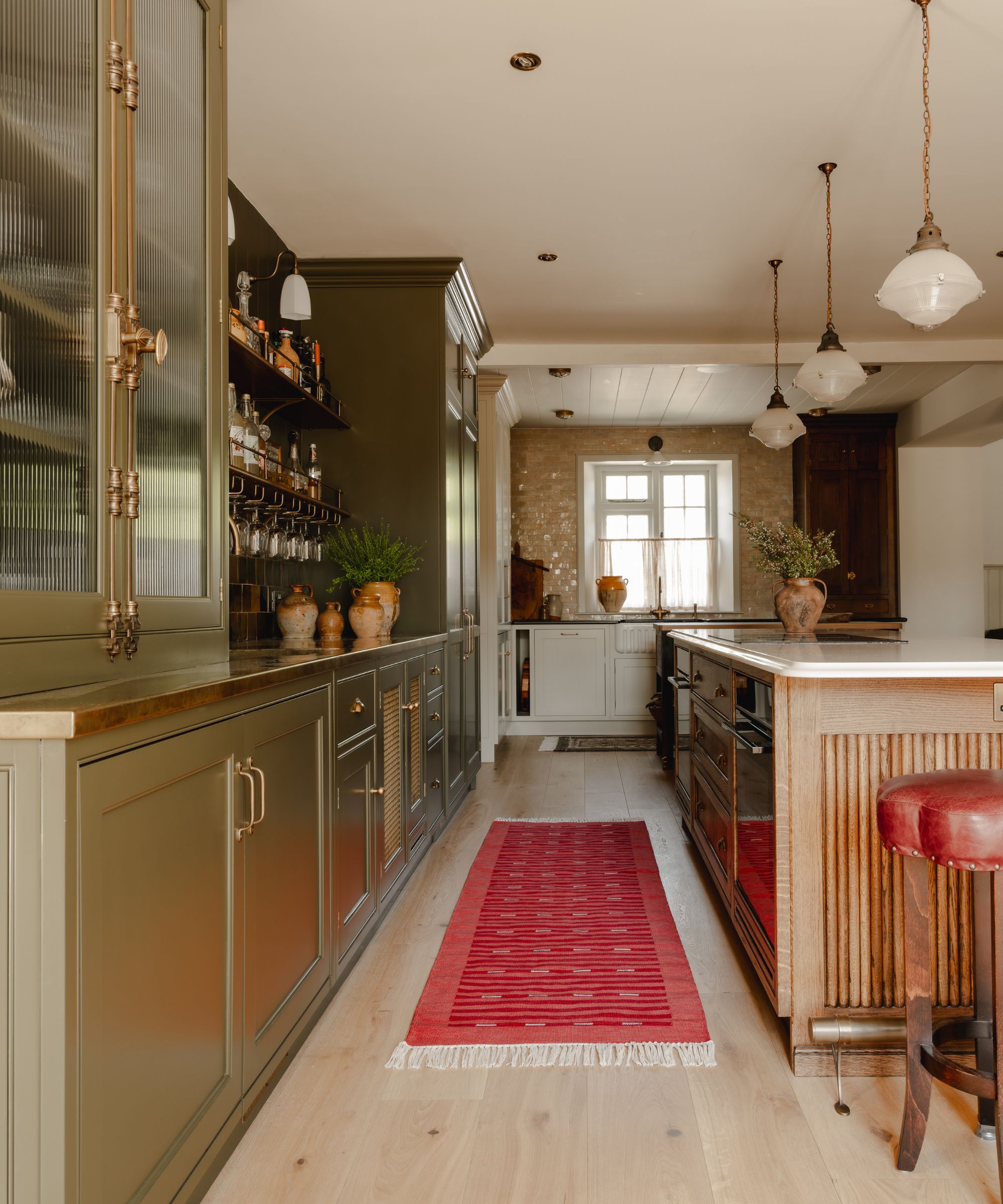 earthy olive green satin painted kitchen with brass hardware, open shelving, and a large wooden island and a red runner rug