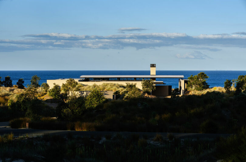A modern, flat-roofed house sits among coastal vegetation, overlooking the ocean under a clear blue sky with scattered clouds.