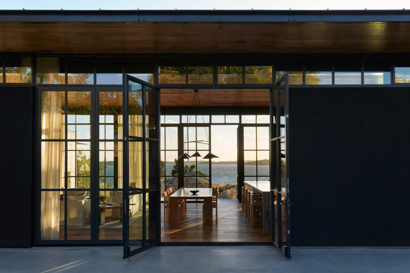 Glass double doors open to a modern dining area with wooden floors, large windows, and views of a lake and distant hills at sunset.