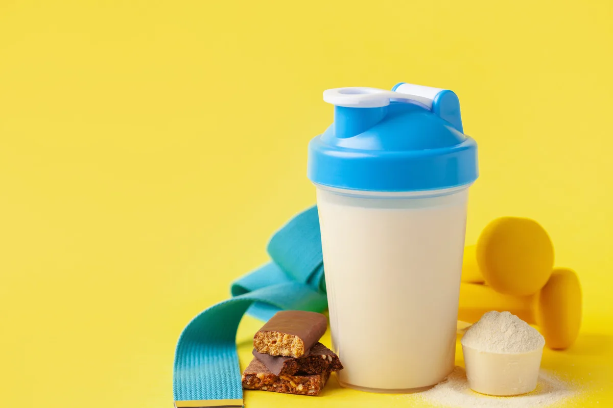 A shaker full of protein powder, a protein bar and a set of weights against a bright background