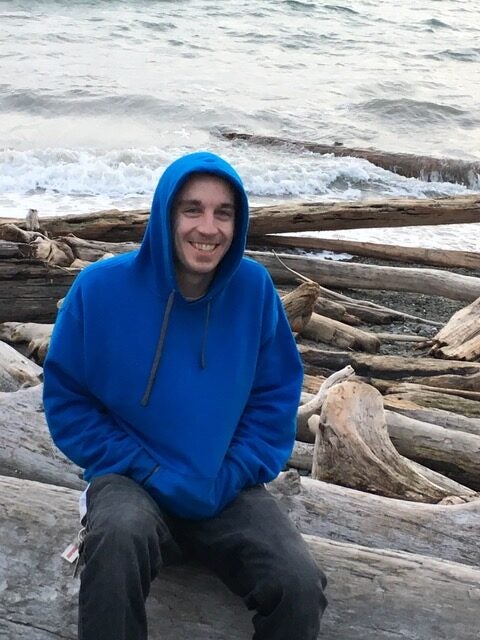 A person in a blue hoodie sits on driftwood logs by the shoreline, with waves and scattered logs in the background.