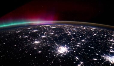 A view of Earth from space at night, showing bright city lights and a colorful aurora with green and pink hues in the atmosphere above the horizon.