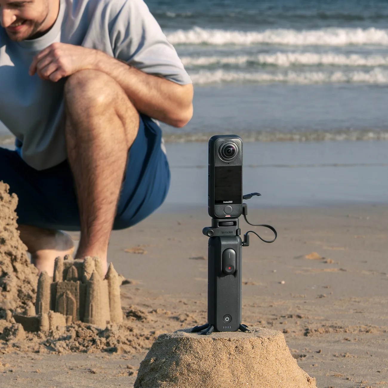 Black Insta360 camera mounted on a small tripod on a sandy beach near a sandcastle.