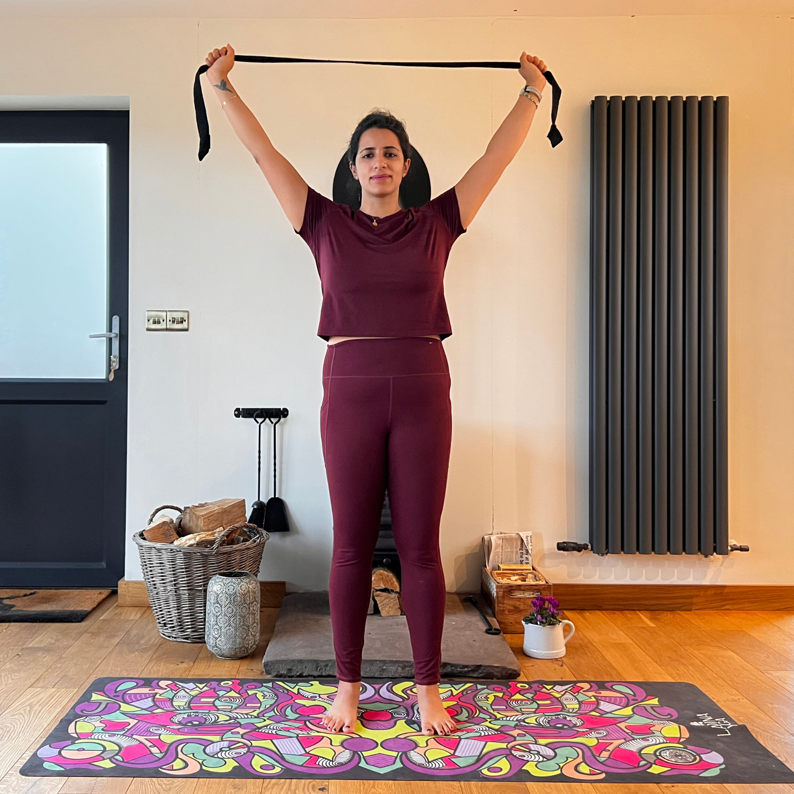 Maysun Hassanaly performs a shoulder floss movement with a yoga strap. She is standing on a colorful yoga mat, holding the strap overhead with wide arms. 