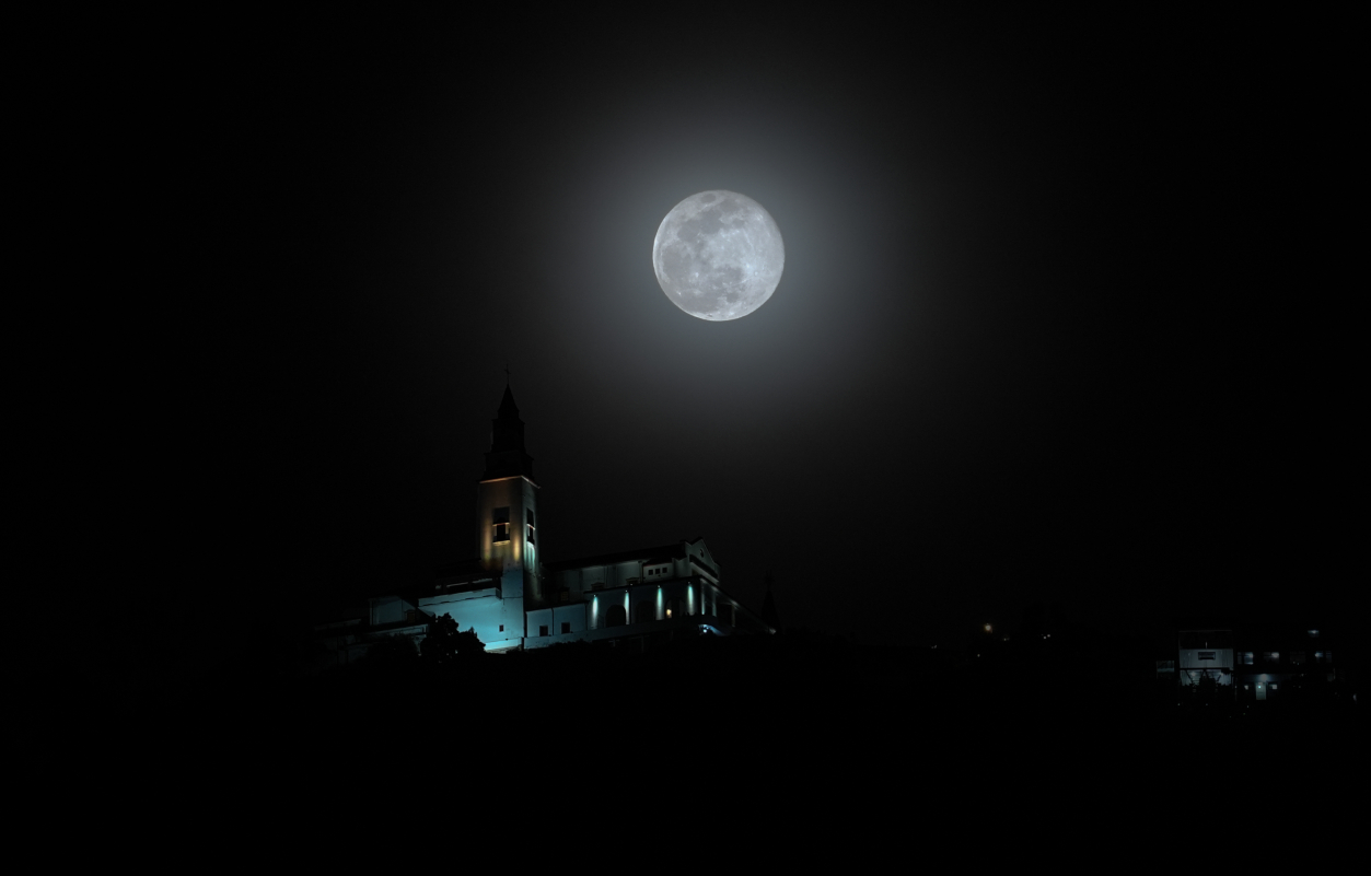 A full moon is surrounded by a ghostly glow in a black sky as it shines above a church-like building, surrounded by shadow.