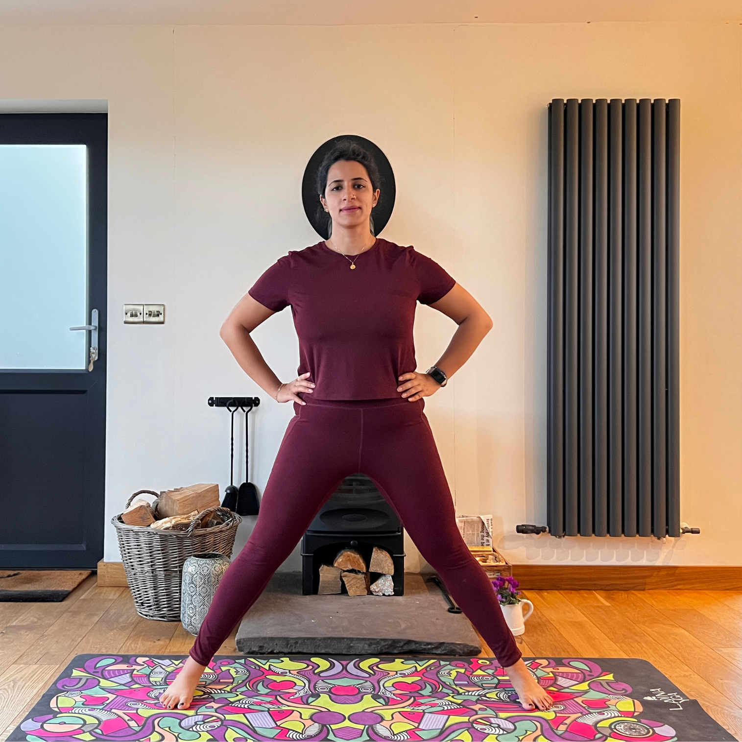 Maysun Hassanaly performs a wide legged forward fold. She is standing on a colorful yoga mat with her legs wide apart and hands on hip.