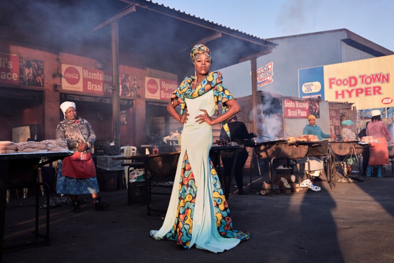 A woman in a colorful, elegant gown poses confidently in an outdoor market, with vendors grilling food in the background and signs for "FOOD TOWN HYPER" and other shops visible behind her.