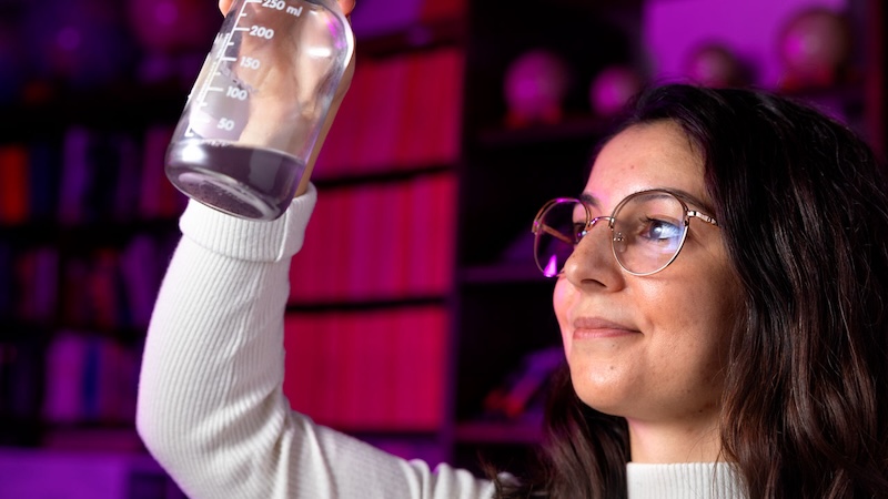 Smiling woman with eyeglasses and dark hair holding a flask with some dark liquid inside it in a laboratory.