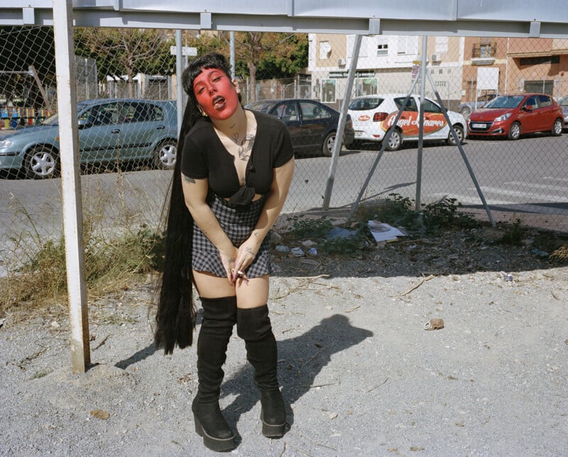 A person with long black hair, dramatic red face paint, black top, plaid skirt, and black thigh-high boots poses outdoors on a gravel lot with parked cars and a chain-link fence in the background.