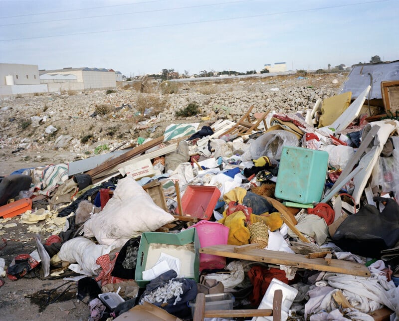 A large pile of assorted garbage and discarded household items is scattered on the ground in an open, barren area with industrial buildings in the background.