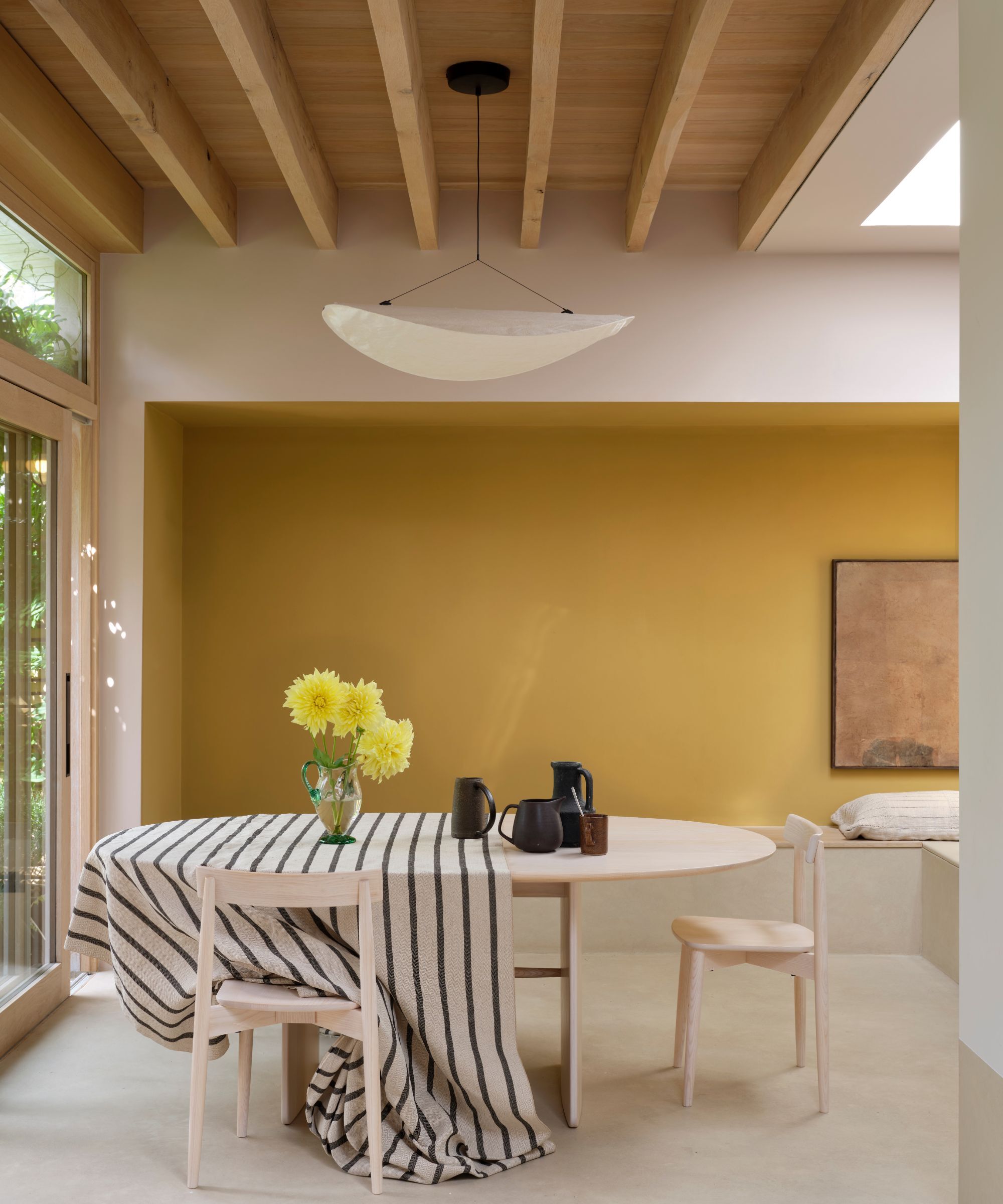 A room with a rear wall painted in a mustard colour, ceilings in a pale creamy pink. There is a large table in the middle of the room with two chairs. There is a striped table cloth on top with a glass jug of yellow dahlias and a few black jugs.