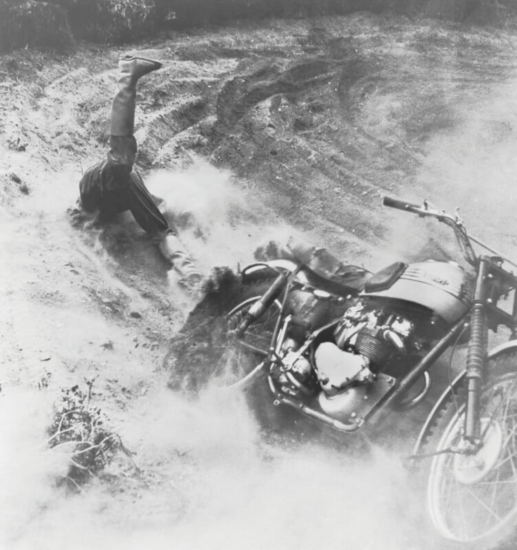 A person has fallen off a motorcycle on a dirt track, with their legs in the air and body on the ground amid a cloud of dust, while the motorcycle lies on its side nearby.