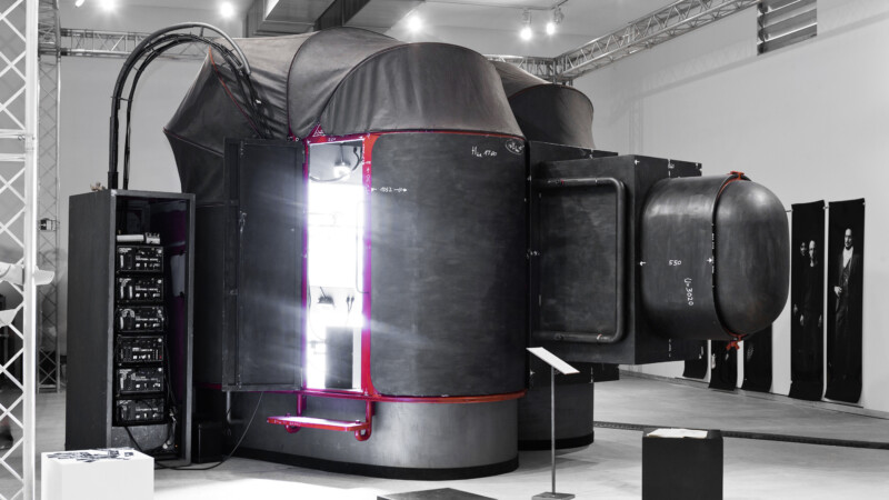 A large, dark, futuristic machine with curved surfaces and protruding cylindrical compartments is displayed in an exhibition space under bright lights. There are open panels revealing technical components inside.