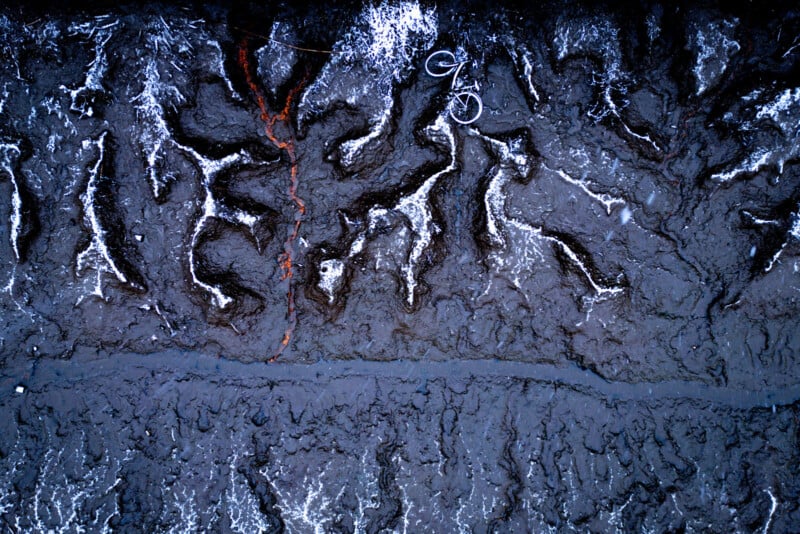 Aerial view of rugged, eroded gray landscape with deep grooves and white accents, featuring a bicycle lying on its side near the top center of the image.