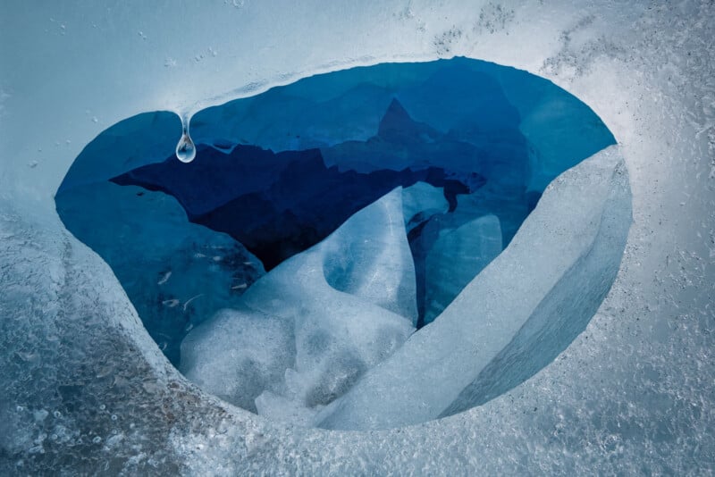 A close-up view of a hole in thick ice reveals deep blue layers and jagged formations underneath, with a single water droplet hanging from the edge of the opening.
