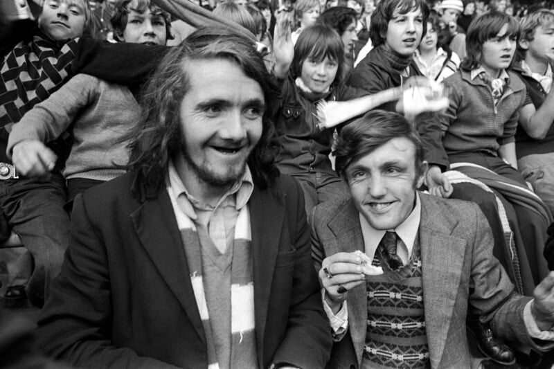 Two men sitting in a lively crowd at a sporting event, smiling and dressed in jackets and scarves. People in the background appear cheerful and animated, suggesting excitement and camaraderie. The image is in black and white.