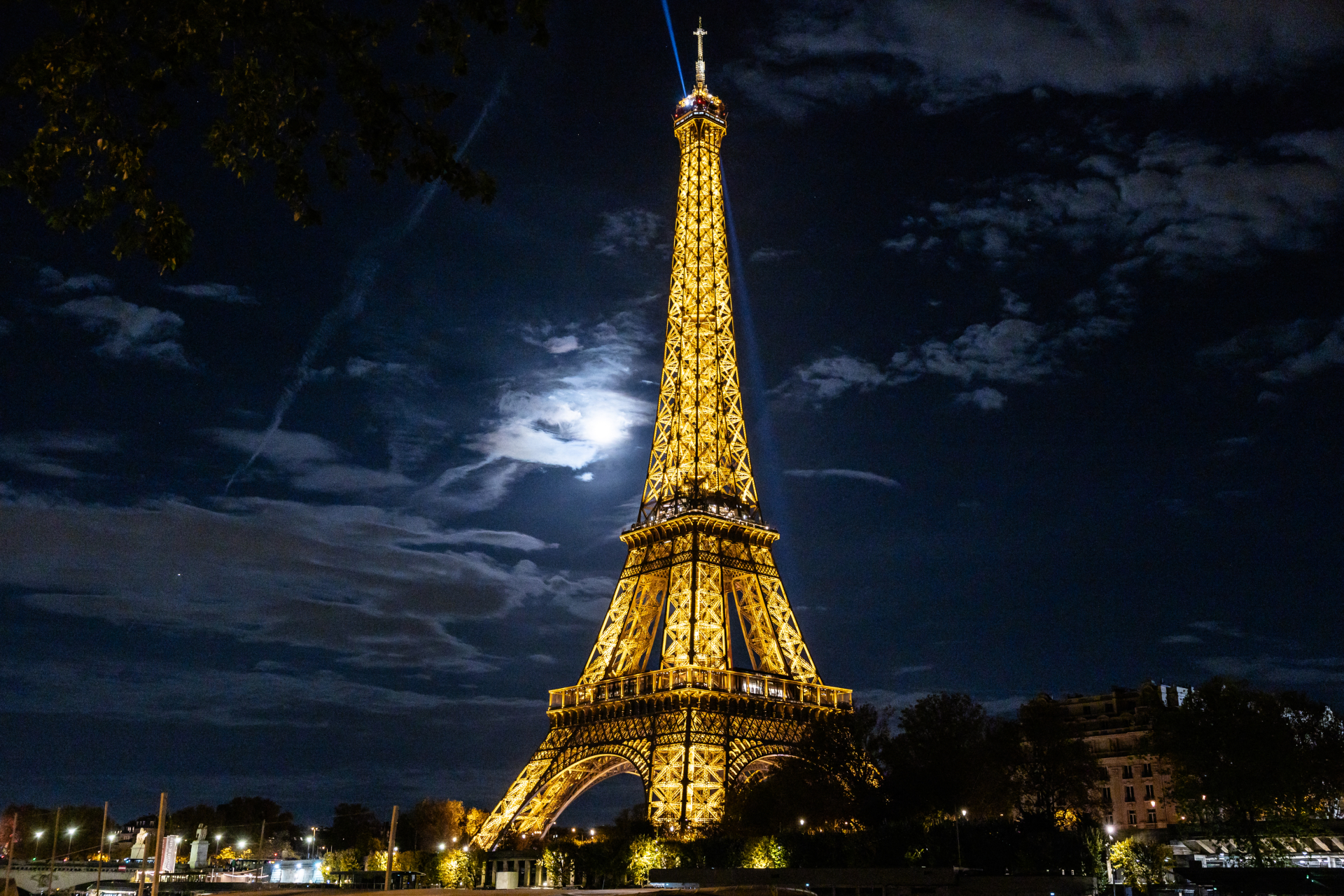 A view looking up at the Eiffel Tower lit in gold at night shining above the Paris skyline as the moon illuminates dappled clouds to the left of the steel structure.