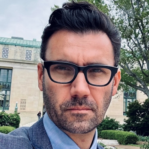 Man with black eyeglasses, dark hair and scruffy beard and mustache, with a building behind him.