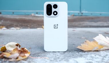 A white OnePlus smartphone with a distinctive rear camera design is placed on the ground surrounded by autumn leaves.
