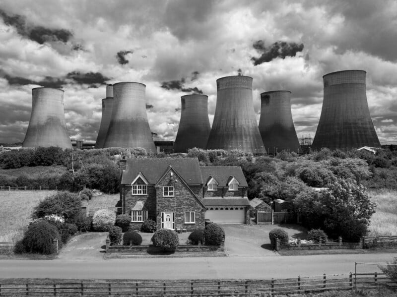 A large brick house with a neatly kept garden stands in front of a row of massive industrial cooling towers under a cloudy sky. Shrubs and fencing separate the property from the imposing structures behind.