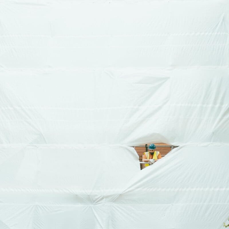 A construction worker in a yellow vest and blue helmet is visible through a tear in a large white plastic sheet that covers a building. The worker is standing behind the sheet, partially obscured.