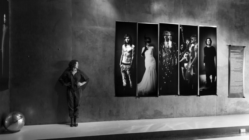 A person stands with hands on hips beside a wall displaying five large portraits of differently dressed individuals. The scene is in black and white, with modern, minimalist decor and a reflective silver ball on the floor to the left.