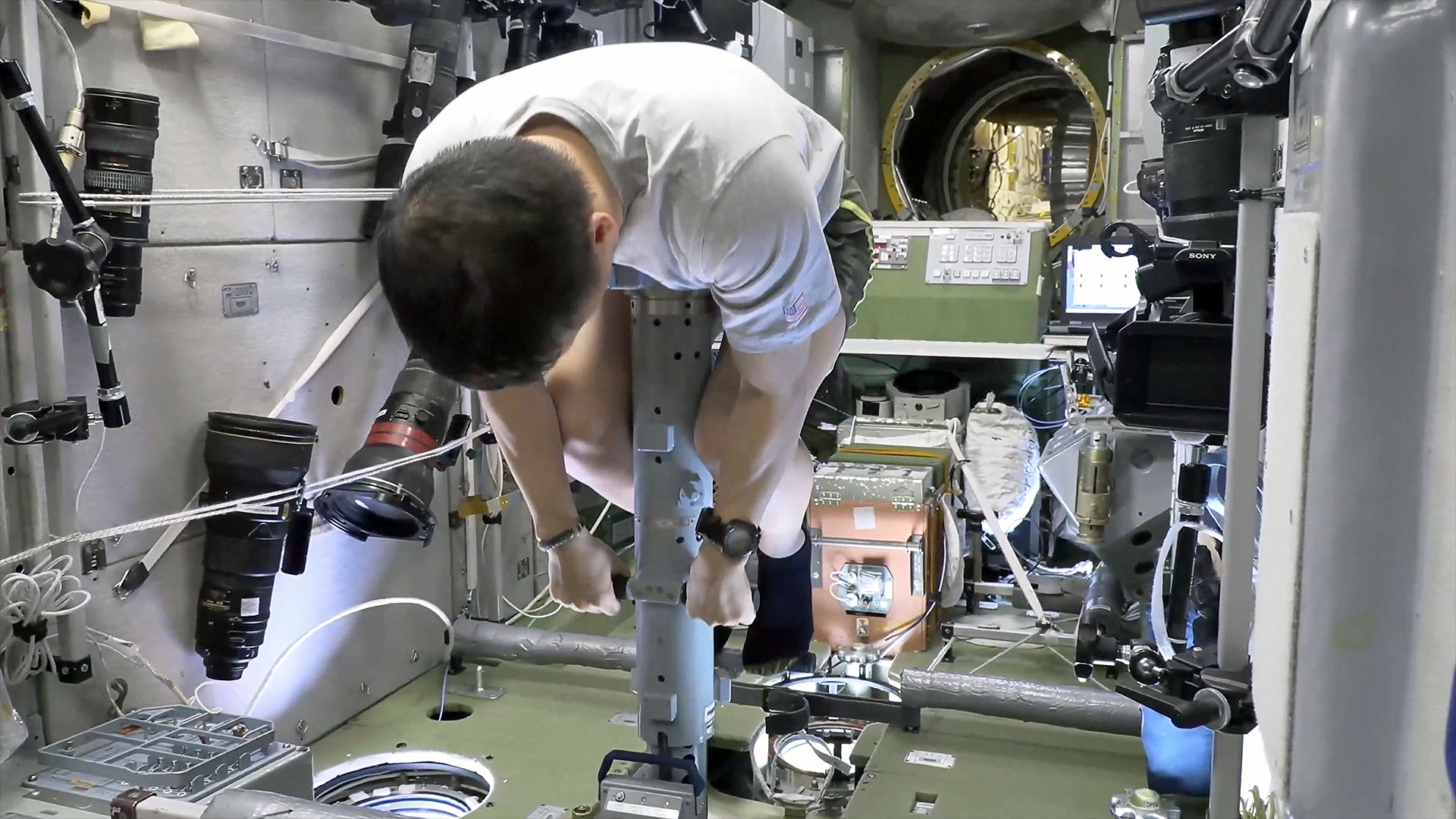 A man with dark hair wearing a grey t-shirt leans over a grey metal cylinder inside a space station