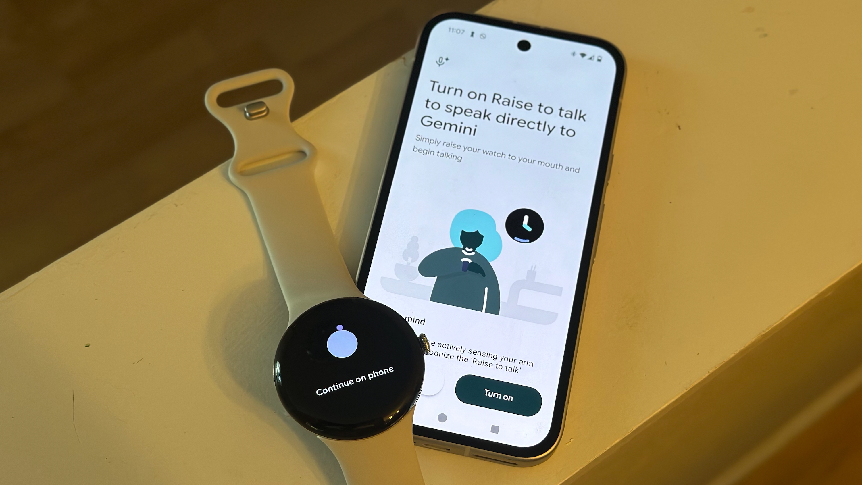 A photo of the Google Pixel Watch 4 sitting atop a Google Pixel 9a smartphone on a shelf, the watch showing "Continue on phone" and the phone showing "Turn on Raise to talk to speak directly to Gemini," with a "Turn on" button visible.
