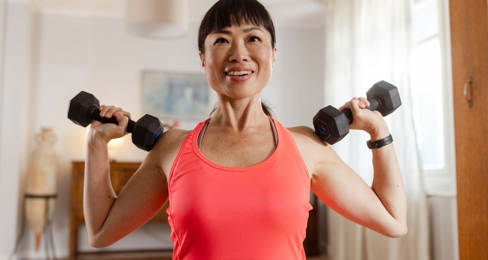 woman facing the camera from the waist up wearing a pink vest holding a dumbbell in each hand at her shoulders, ready to press them up. there's a home setting behind her.