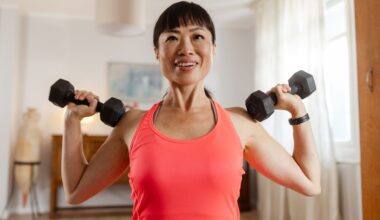 woman facing the camera from the waist up wearing a pink vest holding a dumbbell in each hand at her shoulders, ready to press them up. there's a home setting behind her.