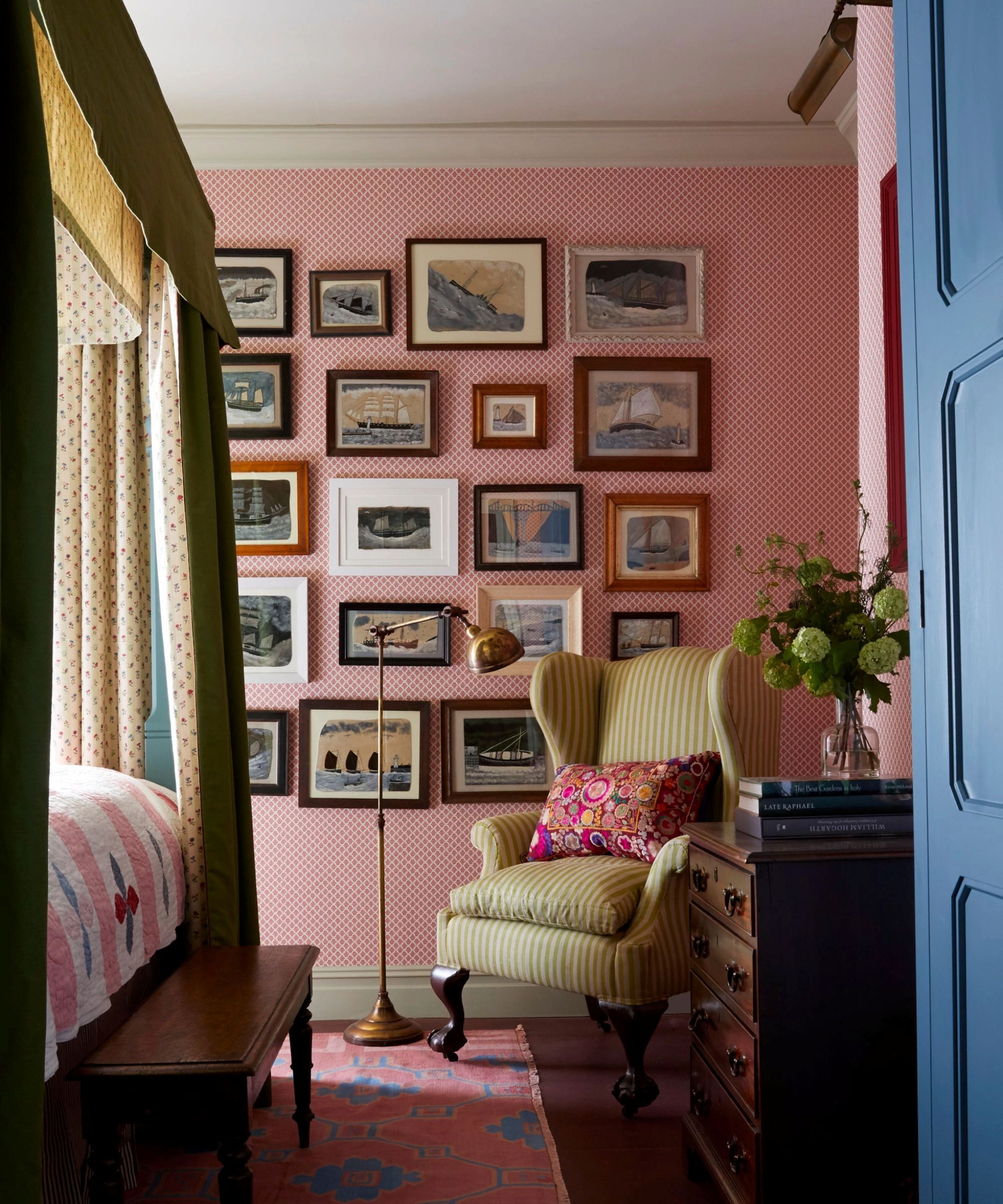 pink bedroom with a gallery wall of sea and boat paintings, a four poster canopy bed with mustard yellow and patterned rapes, a pink and blue rug, yellow armchair and floor lamp