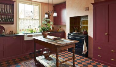 Kitchen with wooden mid-century island, Range Cooker and burgundy built-ins, large window with lace window treatment, open wall-mounted plate rack and orange and navy tiled floor