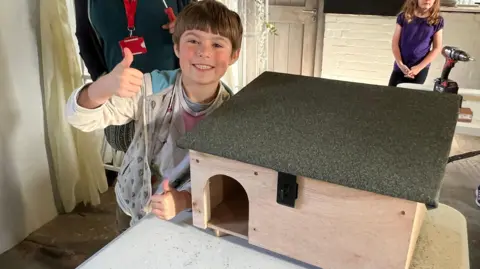Kid smiling and looking at the camera. He's got his thumbs up and smiling next to a hedgehog shelter he's built. It's wooden and has a felt roof on it.
