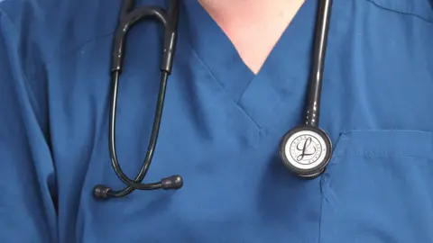 PA Media A person wearing a blue medical uniform and a stethoscope around their neck. The image is cropped in on their upper chest and neck area.