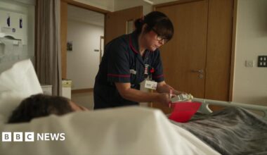 A care staff member at a hospice bed which has a patient in it. She is wearing blue overalls and is looking at medication.
