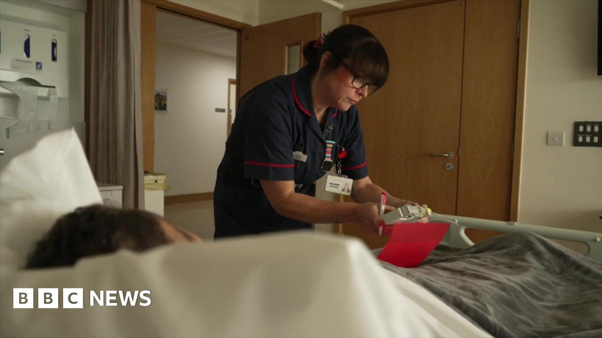 A care staff member at a hospice bed which has a patient in it. She is wearing blue overalls and is looking at medication.