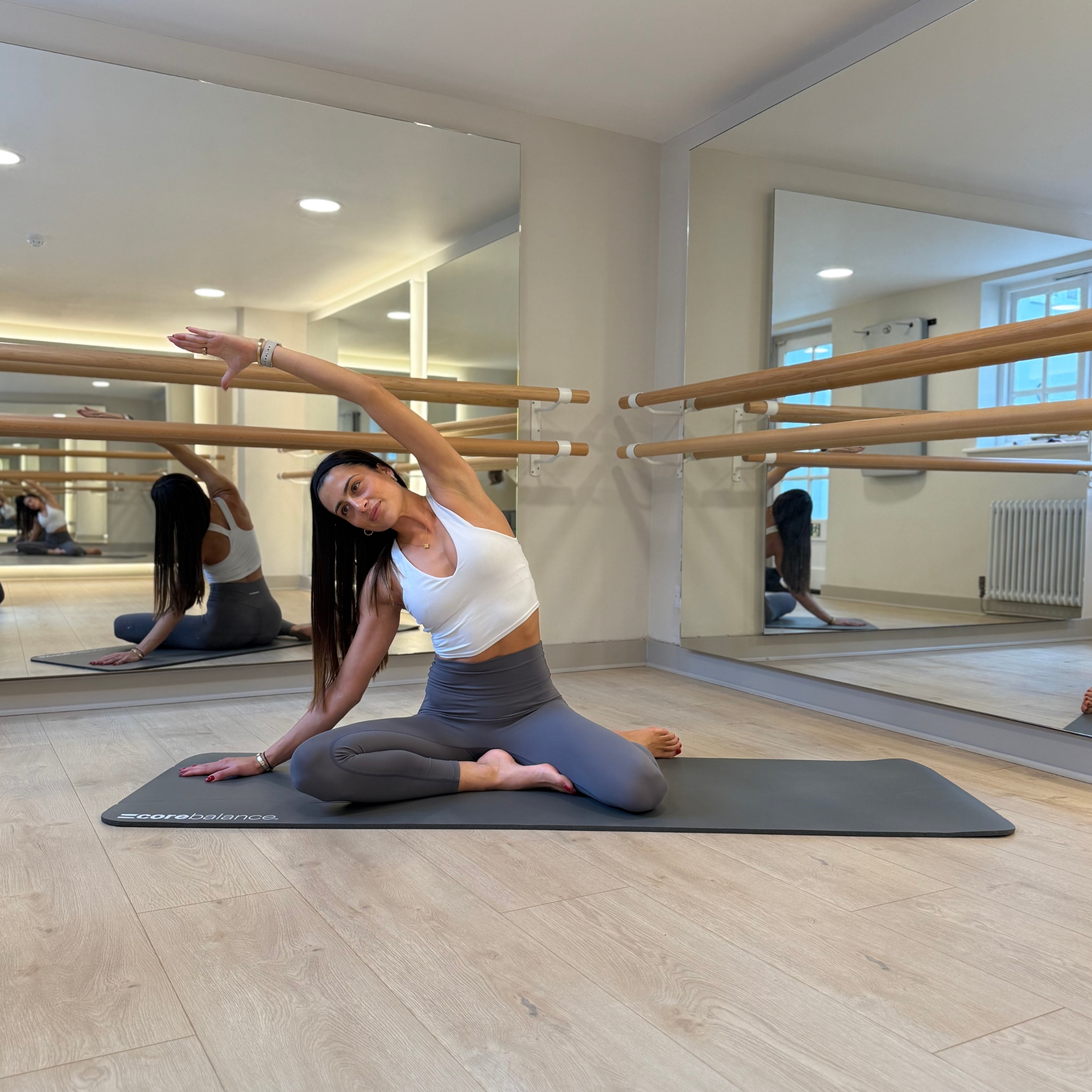 Pilates instructor Mia Gianquitto demonstrates a mermaid stretch in a studio. Her left knee is out to the left side, her left foot pointing backward, her right knee is out to the right side, her right foot resting on her left thigh. She is reaching up and over with her left hand.