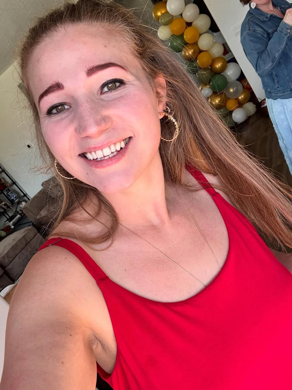 Mykelti Brown smiles for a selfie in a red tank top with hoop earrings, with colorful balloons in the background.