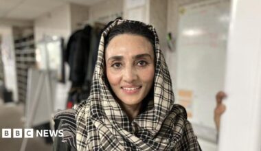 A woman in a black and white checked headscarf smiles at the camera. The background behind is blurred but she is standing in a white room with various objects in it.