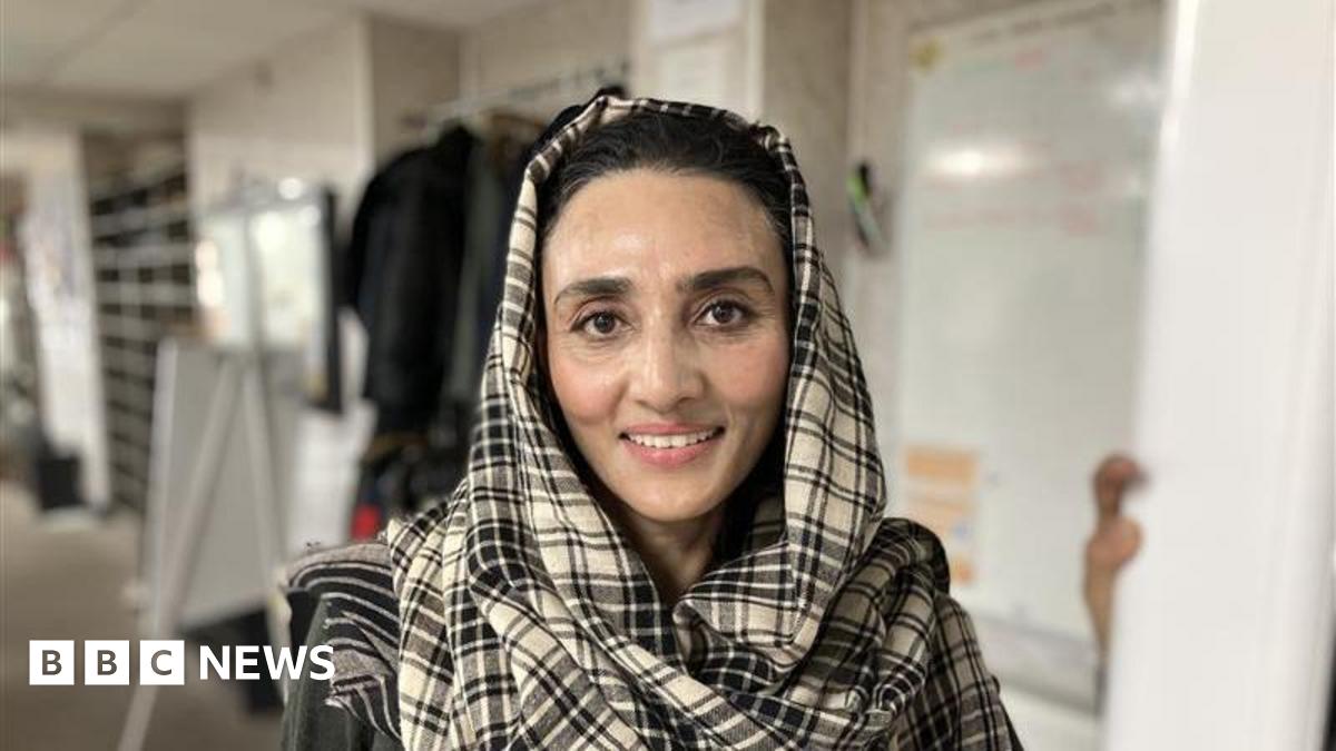 A woman in a black and white checked headscarf smiles at the camera. The background behind is blurred but she is standing in a white room with various objects in it.