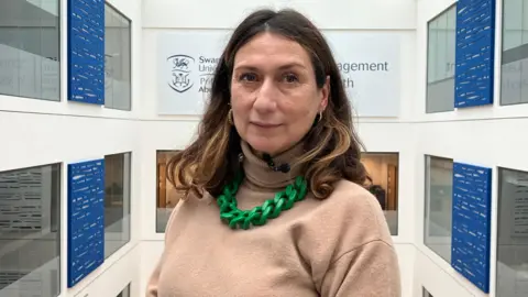 A woman with shoulder length brown hair, wearing a beige turtleneck jumper with a chunky bright green chain necklace looks straight into the camera. Behind her is a series of windows and blue screens on white walls.