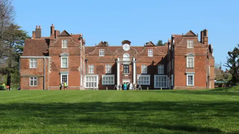 Ipswich Borough Council Christchurch Mansion is in the middle with a stretch of grass in front of it. The building is made of red bricks, and has multiple windows that are painted white. A group of people are standing outside the entrance which is in the middle.
