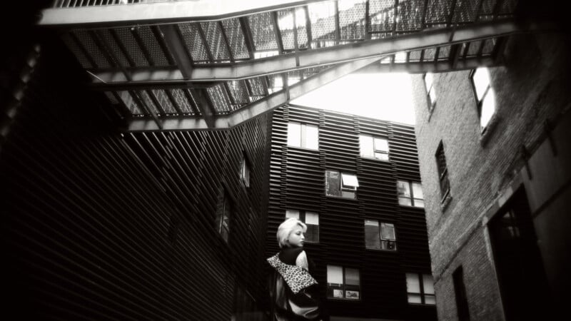A black-and-white photo of a person standing in a narrow alley between modern buildings, looking back over their shoulder. Metal stairs hang overhead, and rectangular windows reflect the light above.