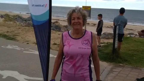 Faye Hussey Faye Hussey smiling at the camera. She is wearing a pink top and is stood in front of a sandy beach. 
