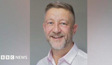 Young 18 year old man sits in the passenger seat of a black VW car, his arm resting on the open window as he looks to camera
